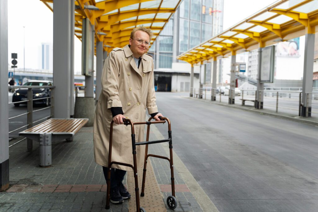 Vyresnio amžiaus žmogus vaikšto su vaikštyne lauke, šypsosi ir mėgaujasi judėjimo laisve.